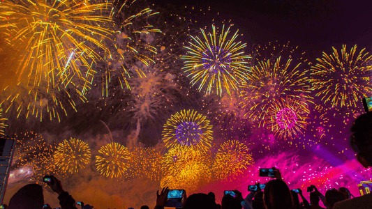 Colorful fireworks bursting in the night sky with silhouettes of people holding smartphones.